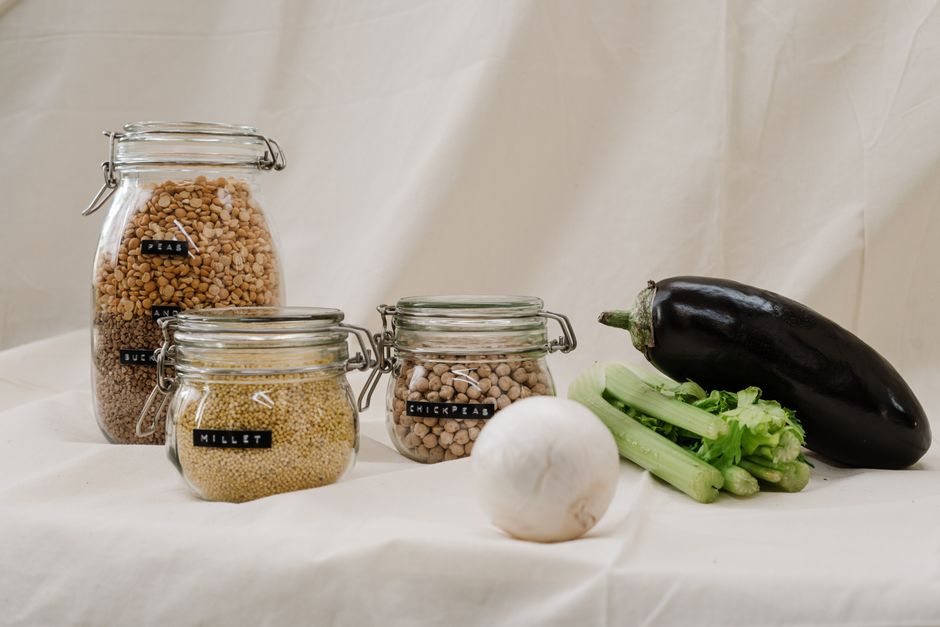 Jars of millet, chickpeas, and vegetables like eggplant and celery on a neutral background.